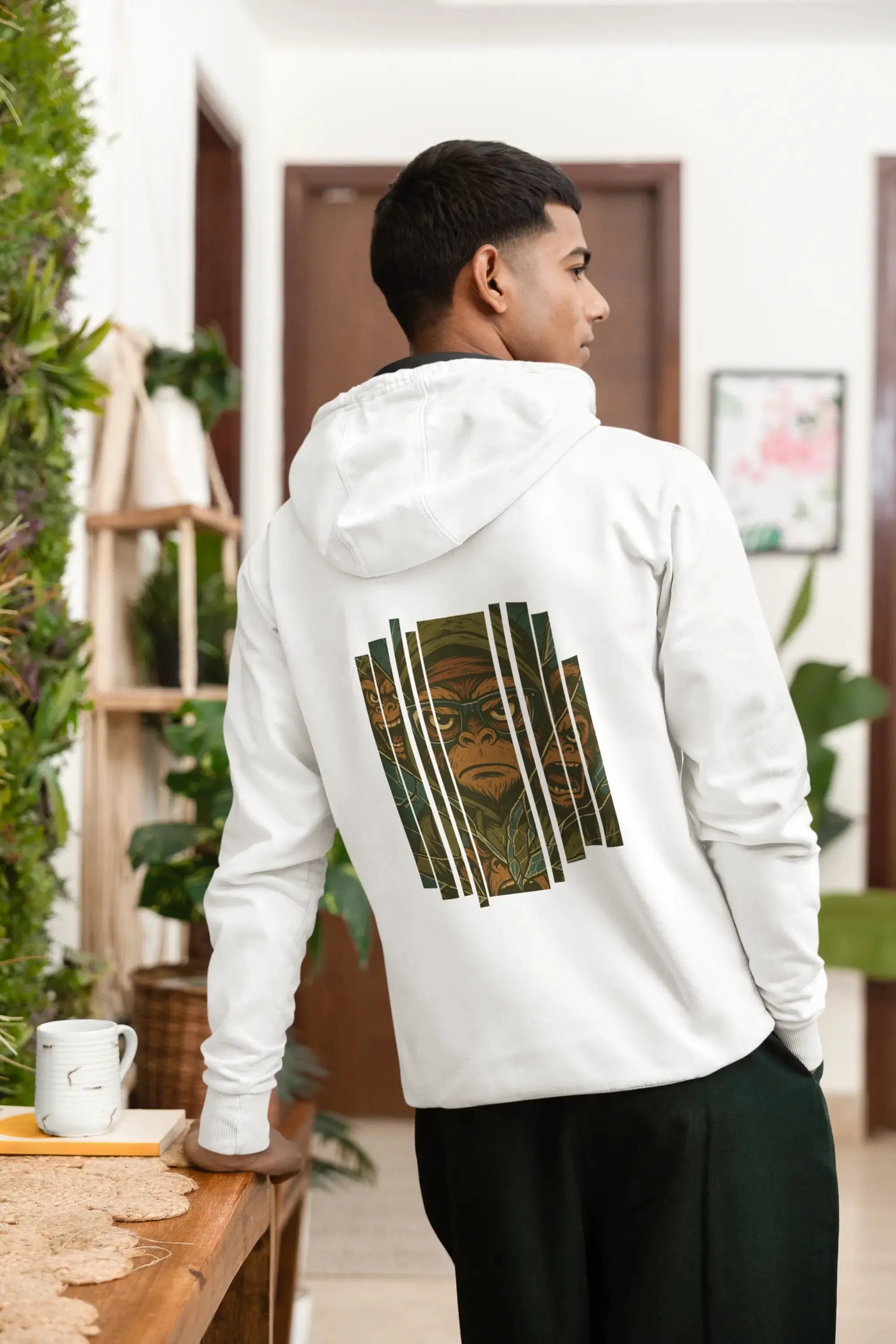 Homme de dos portant un hoodie blanc Artefacts avec une illustration stylisée de Captain Cawette en motif graphique, posé en intérieur près d’une table et de plantes.