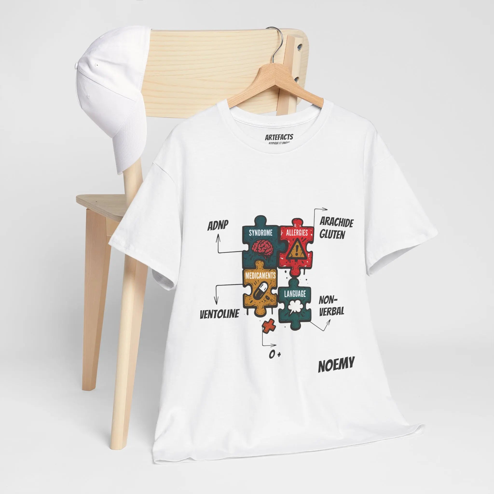 T-shirt blanc personnalisé accroché à une chaise, affichant des pièces de puzzle colorées représentant les informations médicales de Noemy : syndrome ADNP, allergies, médicaments, langage, groupe sanguin et autres éléments essentiels.